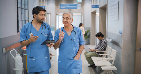 Portrait of Two Indian Male Doctors Walking in Hospital Corridor and Talking while Using a Digital...