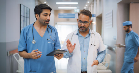 Portrait of Two Indian Male Doctors Walking in Hospital Corridor and Talking while Using a Digital Tablet. Two Medical Specialists Discussing the Treatment of a Patient