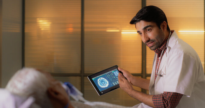 Hospital Ward: Young Indian Male Doctor Talks to Elderly Patient Resting in Bed, Uses Tablet Computer, Shows and Explains Brain CT MRI Scan Results. Physician Talks to a Man Recovering after Surgery