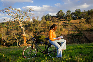 A young black woman reading a book on her bicycle in a field