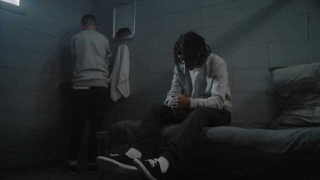 Depressed African American guy sits on the bed in prison cell. Caucasian teenage inmate washes his face in the sink. Young criminals, prisoners serve imprisonment term in jail. Youth detention center.