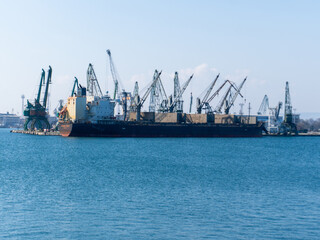 Fototapeta premium Cargo ship and cranes at a busy port, viewed from a cobblestone embankment with seagulls resting