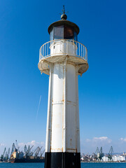 Fototapeta na wymiar Tall white lighthouse with a weathered texture stands against a deep blue sky, overlooking a port with cranes