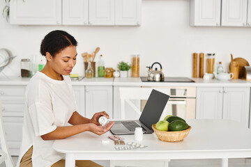 curly african american nutritionist pouring pills into hand palm from medication bottle near laptop