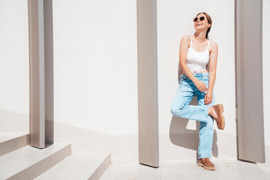 Beautiful smiling model in sunglasses. Female dressed in summer hipster white T-shirt and jeans. Posing near white wall in the street. Funny and positive woman having fun outdoors, in sunglasses