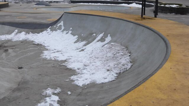 Deserted skate park painted in vibrant yellow and gray hues, within a wintry park landscape, partly covered with remnants of snow on overcast day, without people, captured with a right-to-left pan