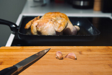 A chicken is being cooked in a pan on a stove. A knife and garlic on a cutting board next to the pan