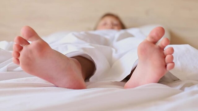 Little girl's feet stick out from under a white blanket at the morning. 