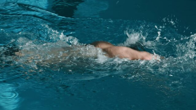 Medium tracking slowmo shot of muscular sportsman in black trunks, rubber cap and goggles swimming front crawl style in lane in pool, while getting ready for triathlon competition