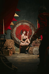 Young redhead woman with makeup and costume sitting and smoking near target over dark retro circus...