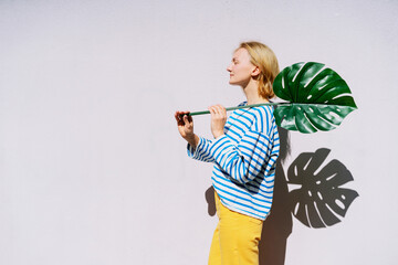Thoughtful woman holding monstera leaf in front of wall. Concept of Ecology, Individuality, Idea