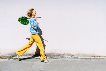 Joyful woman jumping with monstera leaf in hands in front of wall.