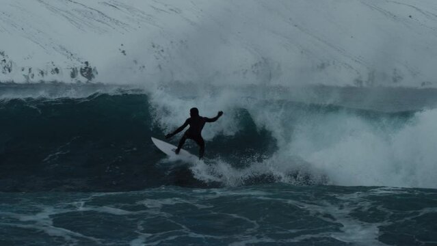 Surfer drops in to bottom turn cutting back and spraying water surfing in arctic with snow on mountains