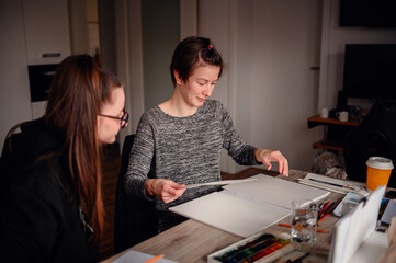 Two women share a creative moment; one chooses paper while the other prepares to paint, a symbol of...