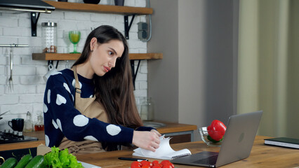 Woman housewife in beige apron tells teacher chef search culinary recipe in notebook study online remote video call conference webcam laptop computer, distance education webinar course in home kitchen