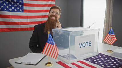 Smiling bearded man with glasses standing by a ballot box in a us voting station, american flags in...