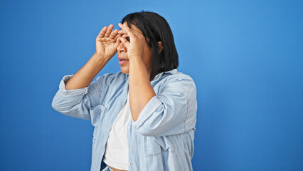 A middle-aged hispanic woman stands against a blue background, peering out with her hands.