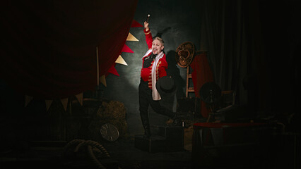 Emotional man with pale face makeup, in red stage costume and hat making performance over dark retro circus backstage background. Concept of circus, theater, performance, show, retro and vintage © master1305