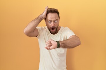 Middle age man with beard standing over yellow background looking at the watch time worried, afraid...