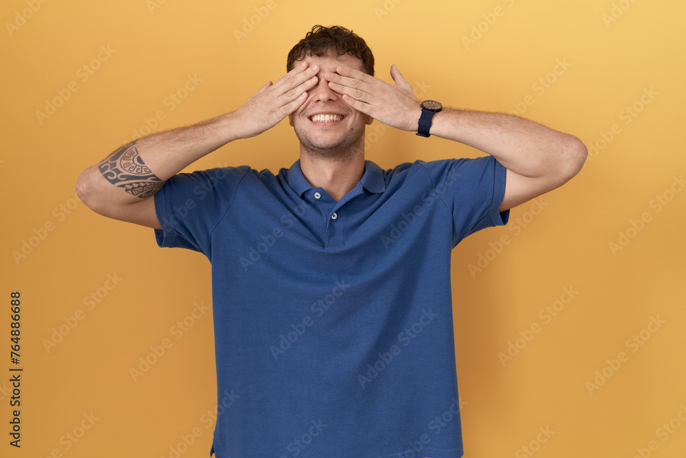 Poster Young hispanic man standing over yellow background covering eyes with hands smiling cheerful and funny. blind concept.