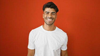 Handsome young hispanic man with a beard smiling against a red isolated background