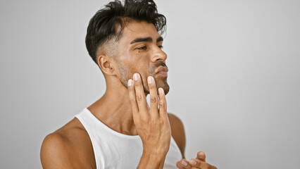 A young hispanic man with a beard touches his face against a white background, portraying grooming.