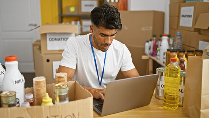 A focused hispanic man with a beard volunteers at a food donation center, managing inventory on a laptop. - obrazy, fototapety, plakaty