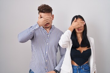 Young hispanic couple standing over white background covering eyes with hand, looking serious and...