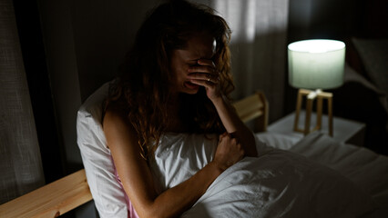 A distraught caucasian woman sitting in bed with lamp indoors at night, portraying stress or...