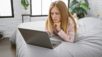 Naklejka premium Young caucasian woman with blonde hair intently using a laptop while lying on a bed in a bright modern bedroom