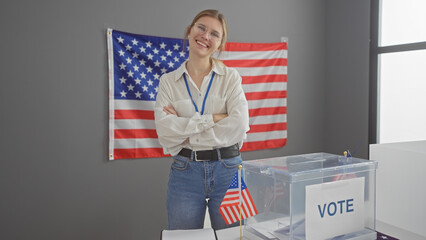 A smiling young woman with crossed arms stands in a voting center with a usa flag behind her, symbolizing democracy.