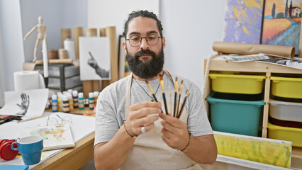 A bearded man examines paintbrushes in an art studio surrounded by paintings, canvases, and art...