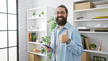 Smiling hispanic man with beard in casual clothing using smartphone gives thumbs up in a modern...