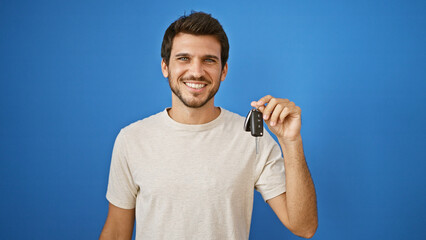 A young hispanic man holding car keys stands smiling against an isolated blue background outdoor.