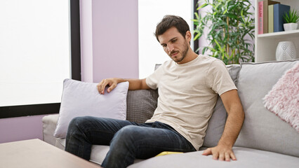 A handsome young hispanic man with a beard relaxes thoughtfully in a modern living room,...