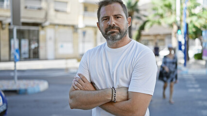 Handsome mature hispanic man with crossed arms standing confidently on a sunny urban street.