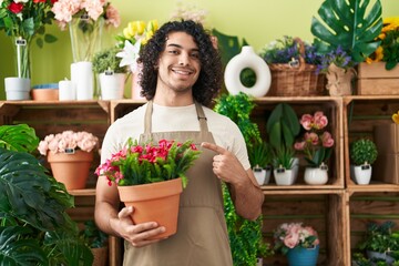 Hispanic man with curly hair working at florist shop holding plant smiling happy pointing with hand and finger