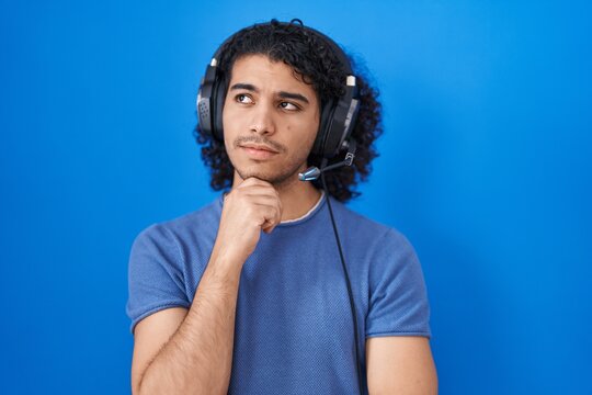 Hispanic man with curly hair listening to music using headphones with hand on chin thinking about question, pensive expression. smiling with thoughtful face. doubt concept.