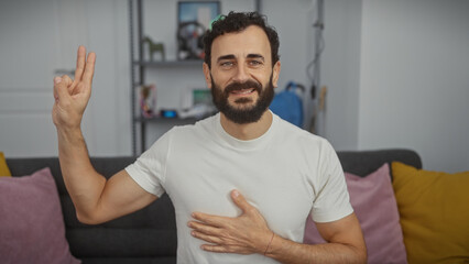 Handsome bearded middle-aged man in a white shirt makes a promise gesture in a cozy living room...
