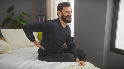 A bearded man in casual attire grimaces with back pain while sitting on a white bed in a modern bedroom interior. - obrazy, fototapety, plakaty