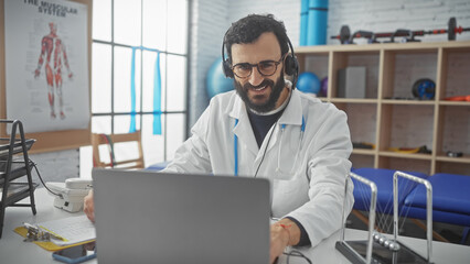 Fototapeta na wymiar Smiling bearded man wearing labcoat and headphones working on laptop in physical therapy clinic