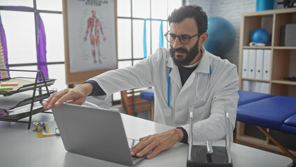 Fototapeta premium A bearded mature man in a white lab coat works on a laptop in a hospital room, exuding professionalism and concentration.
