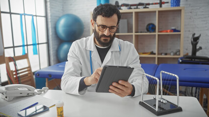 Fototapeta premium Middle-aged bearded man in lab coat using tablet at rehabilitation center clinic.