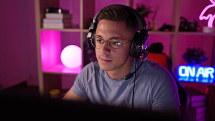 A young hispanic man engages in gaming at night in a dark room illuminated by colorful led lights, wearing headphones and glasses.