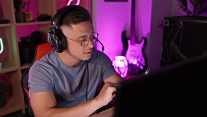 Fototapeta na wymiar A focused young man using headphones works attentively in a dimly lit home office with gaming setup and decor.