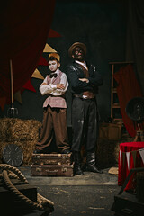 Retro-themed event. High African man standing with little servant boy over dark retro circus backstage background. Concept of circus, theater, performance, show, retro and vintage
