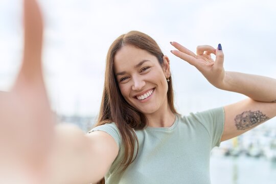 Young beautiful woman making selfie by camera doing victory gesture with fingers at seaside