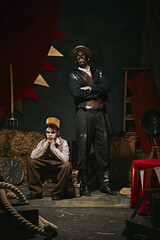 High African man with dirty face standing with little servant boy over dark retro circus backstage...