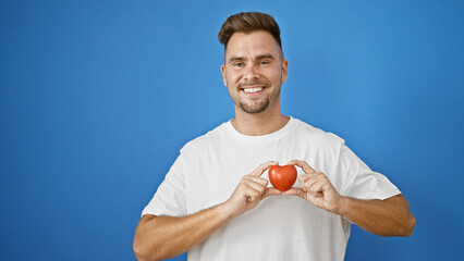 A handsome hispanic man holds a red heart symbol against a plain blue outdoor background,...