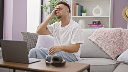 A stressed young man with a beard feeling headache at home in a living room with a laptop.